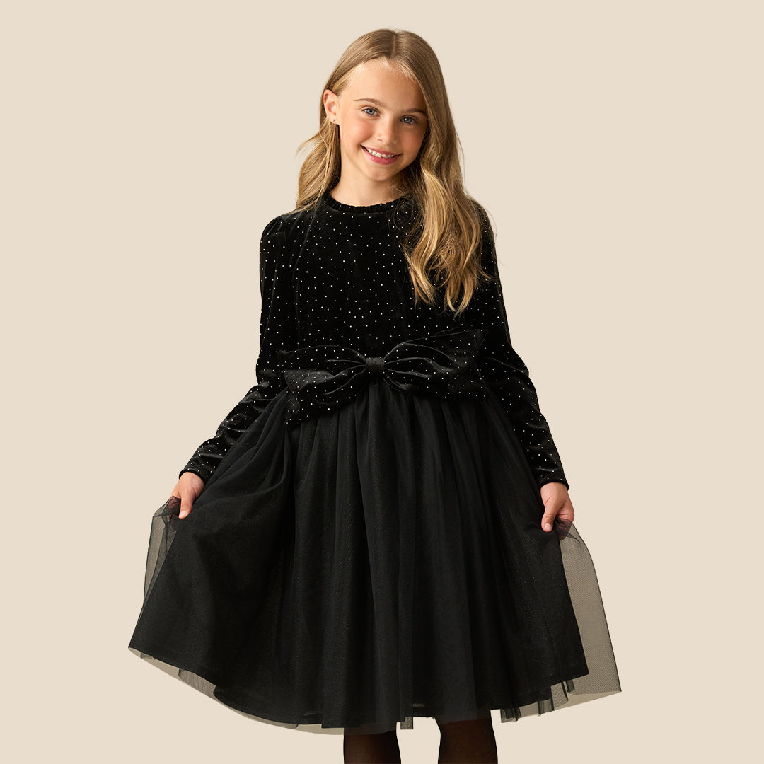 A young girl with blonde hair wearing a black long-sleeve dress with a polka-dot velvet bodice, a sheer black tulle skirt, and a matching bow belt, posing against a solid beige background.