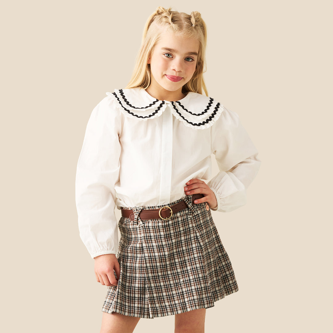 A model wearing a pleated plaid mini skirt in beige, brown, and black, paired with a white blouse and a brown belt with a gold ring, set against a solid beige background.