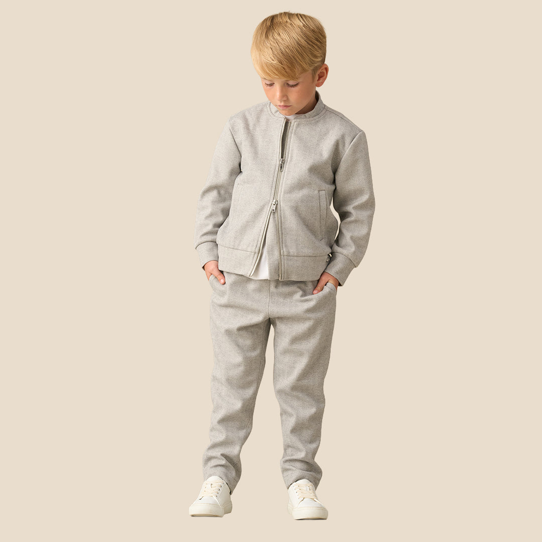 A child wearing light gray heathered sweatpants with a white t-shirt and white sneakers, standing against a solid beige background.