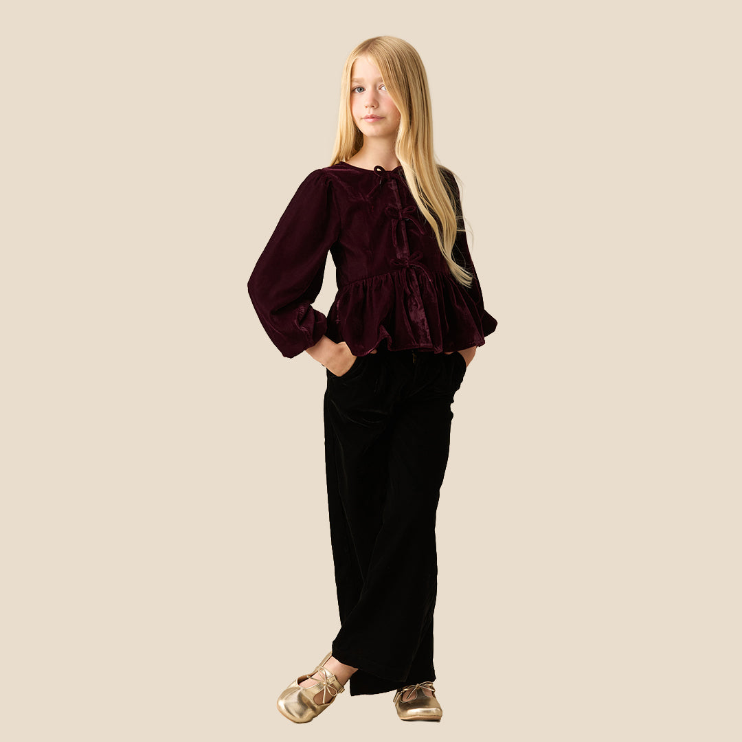 A young girl with long blonde hair wearing a deep burgundy velvet long-sleeve top with a tie-neck and peplum hem, paired with black pants, set against a solid light beige background.