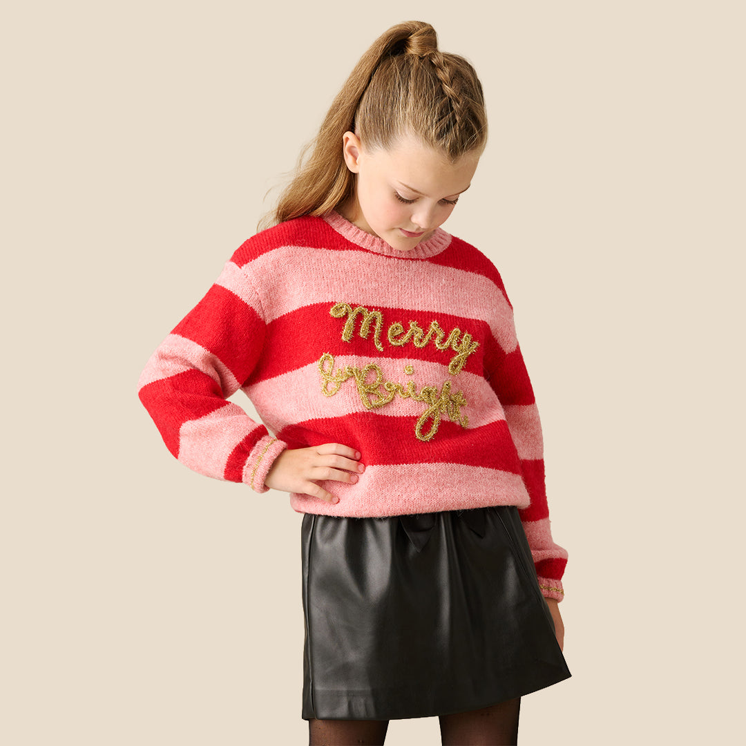 A young girl wearing a fuzzy knit sweater with red and pink horizontal stripes, featuring 'Merry Night' in gold glitter script across the chest, paired with a black faux-leather mini skirt, set against a solid beige background.