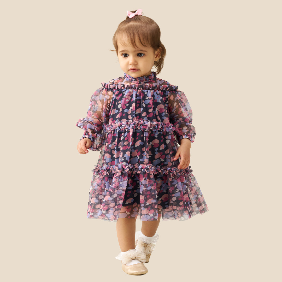 A toddler girl wearing a tiered long-sleeve dress with a dark navy base and a colorful floral pattern in pink, purple, and blue, standing against a solid beige background.
