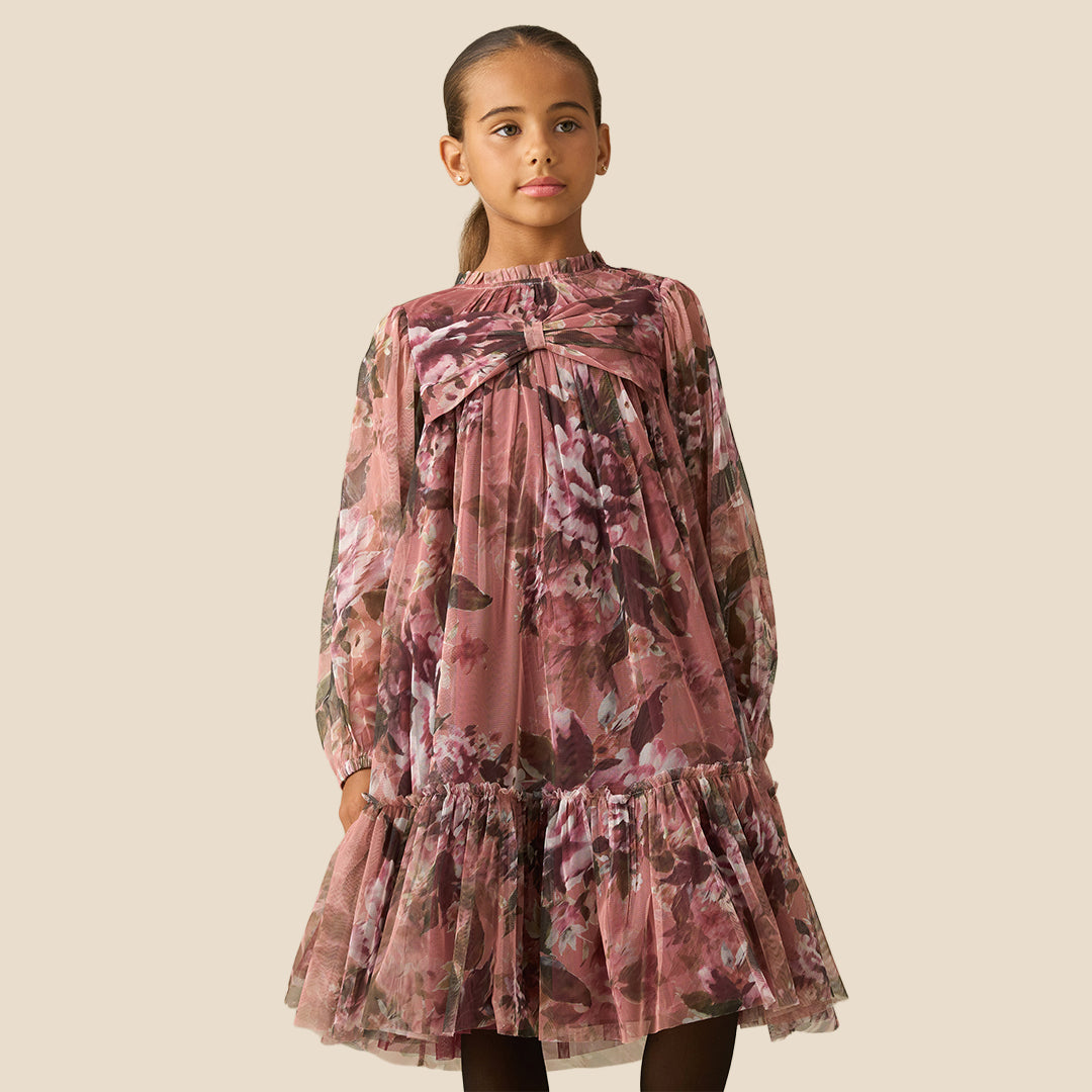 A young girl with her hair in a ponytail models a long-sleeve tiered dress with a dusty rose and brown floral print against a solid beige background.