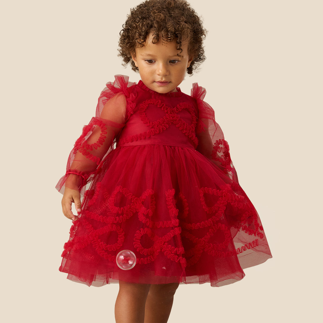 A toddler with curly brown hair wearing a red tulle dress with sheer long sleeves and red floral applique details, standing against a solid beige background.