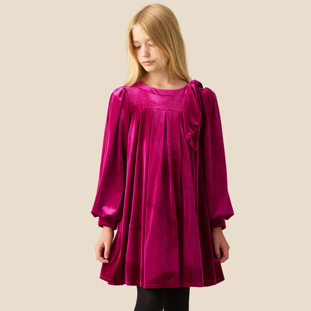 A young girl with blonde hair wearing a long-sleeved magenta velvet dress with a gathered bow detail on the chest, standing against a solid beige background.