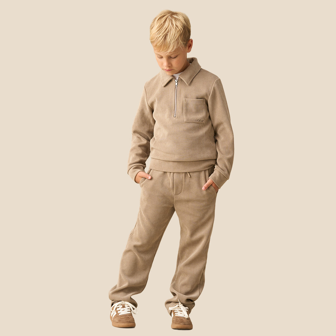 A child wearing a matching beige fleece tracksuit with a half-zip sweatshirt and relaxed-fit pants, paired with brown sneakers, against a solid light beige background.