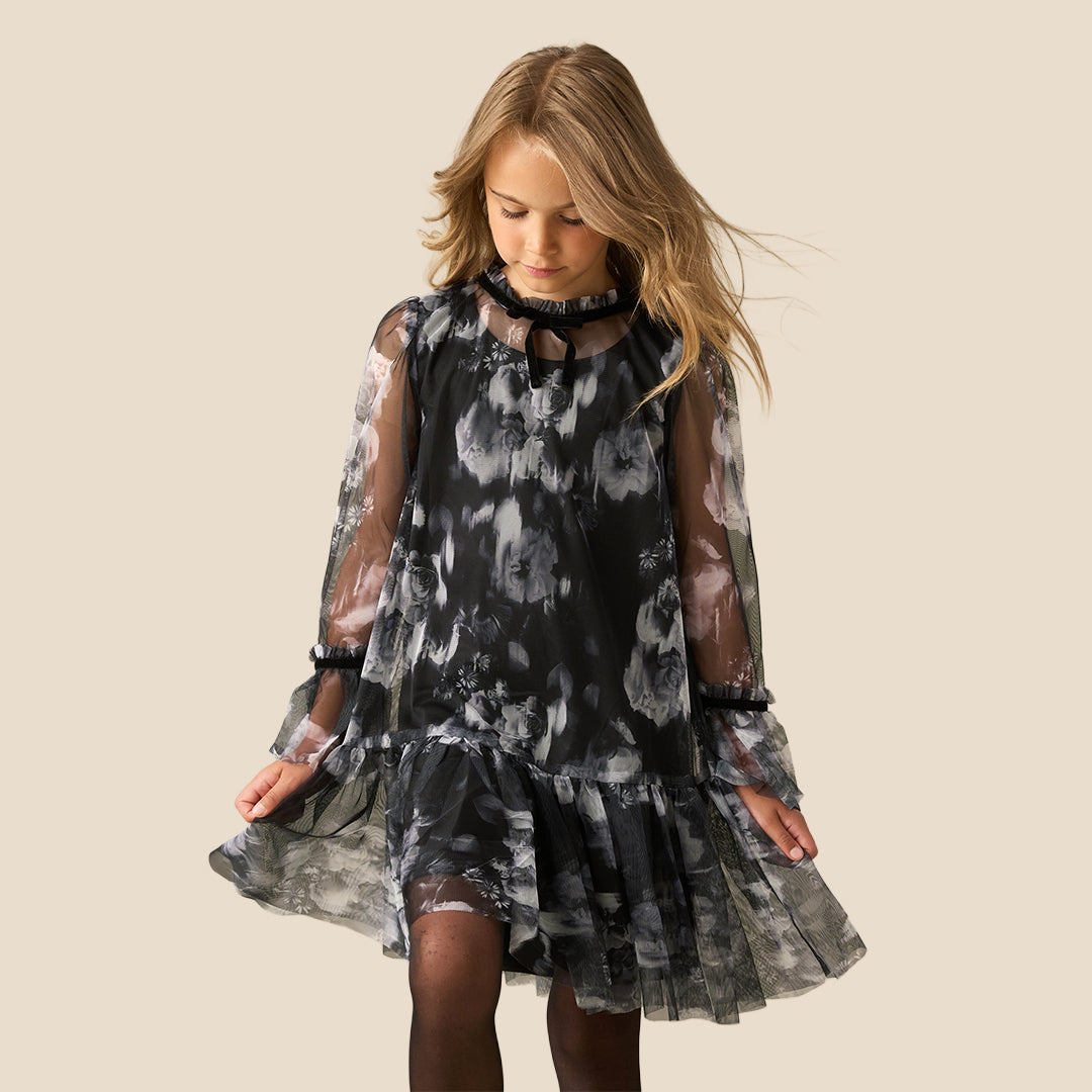 A young girl with long light brown hair is wearing a black and gray tie-dye dress with sheer long sleeves and a ruffled skirt, standing against a solid beige background.