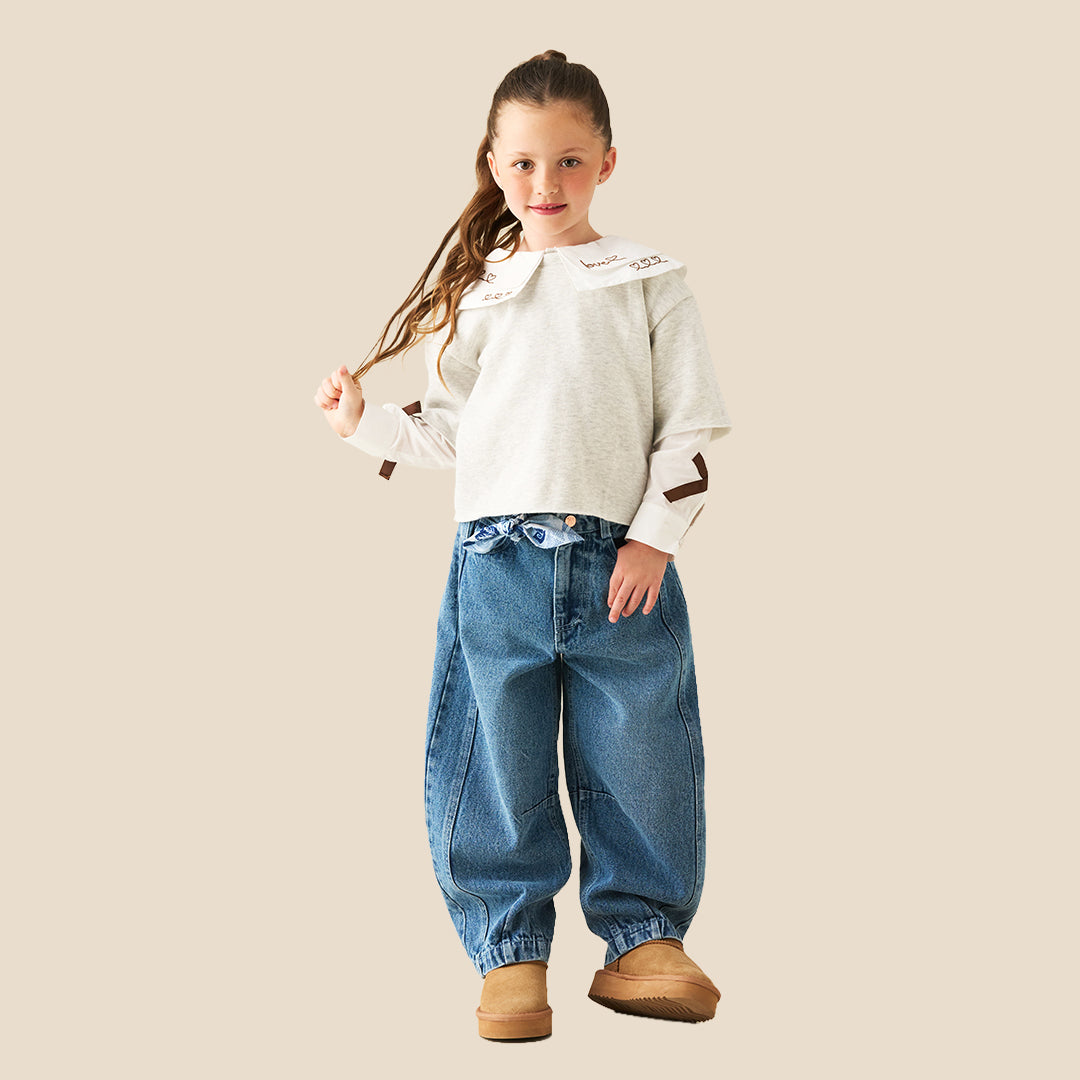 A young girl with her hair in a ponytail models a light gray long-sleeve top featuring a large white collar with brown embroidered details and matching brown accents on the sleeves, paired with blue jeans against a solid beige background.