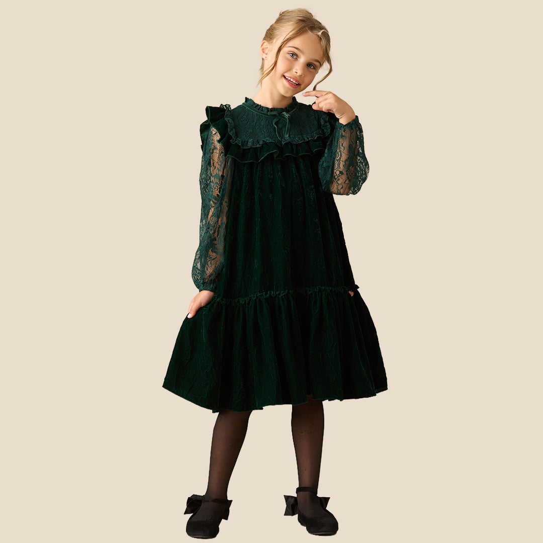 A young girl wearing a dark forest green velvet dress with lace sleeves and a ruffled high neckline, photographed against a solid beige background.