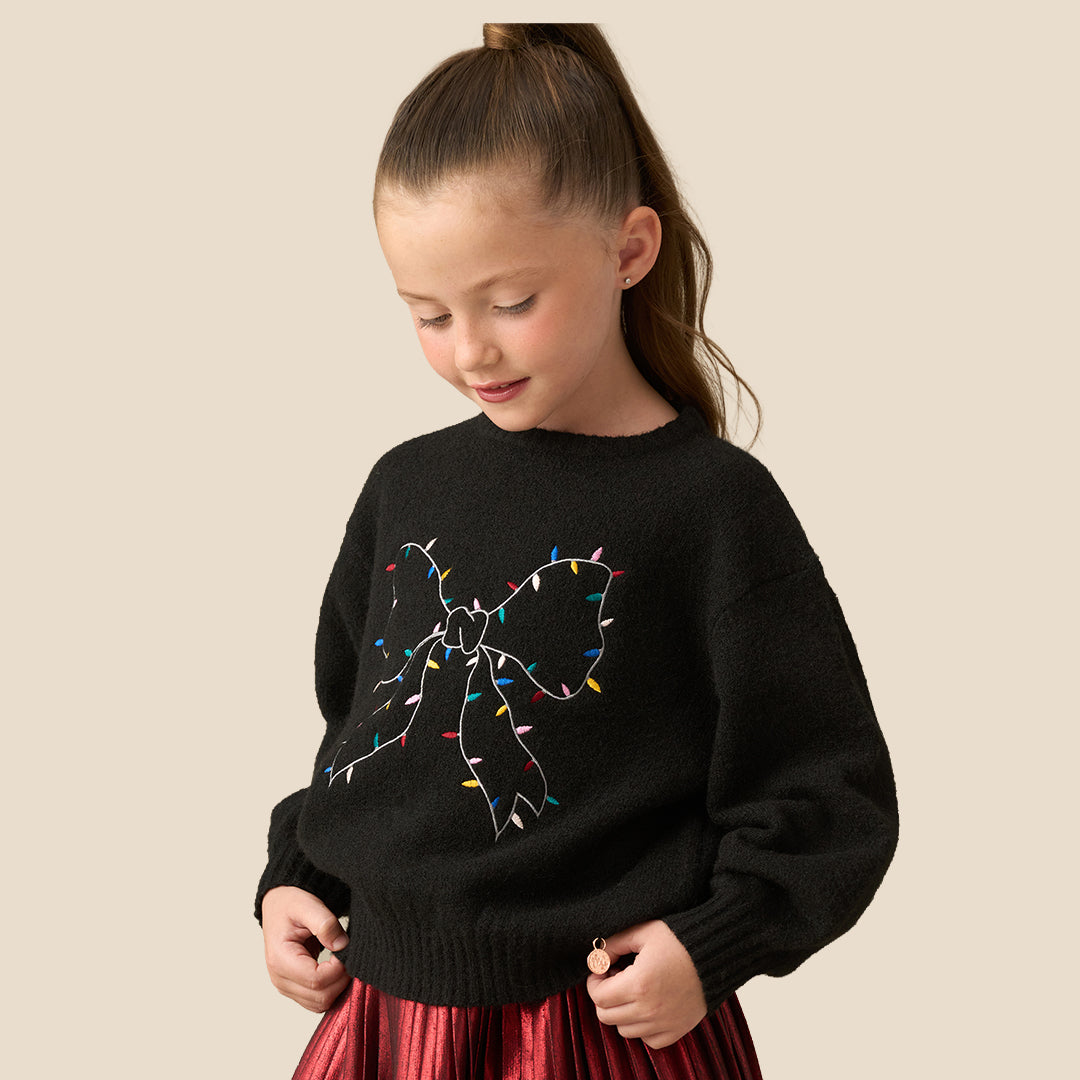 A young girl with her hair in a ponytail is wearing a black knit sweater featuring a large bow design outlined with colorful Christmas string lights, paired with a red pleated skirt, set against a solid beige background.