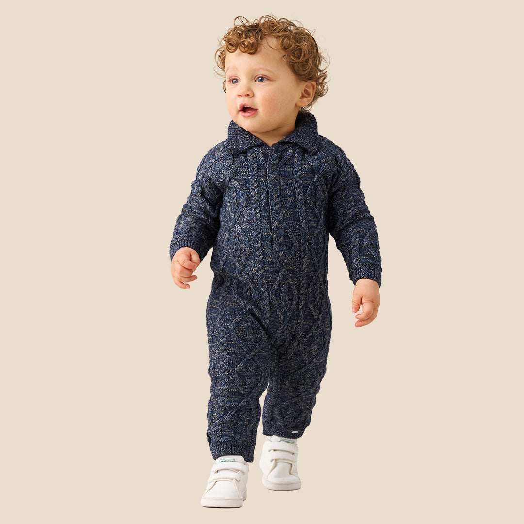 A baby with curly light brown hair wearing a navy blue cable-knit one-piece jumpsuit and white shoes, standing against a solid beige background.