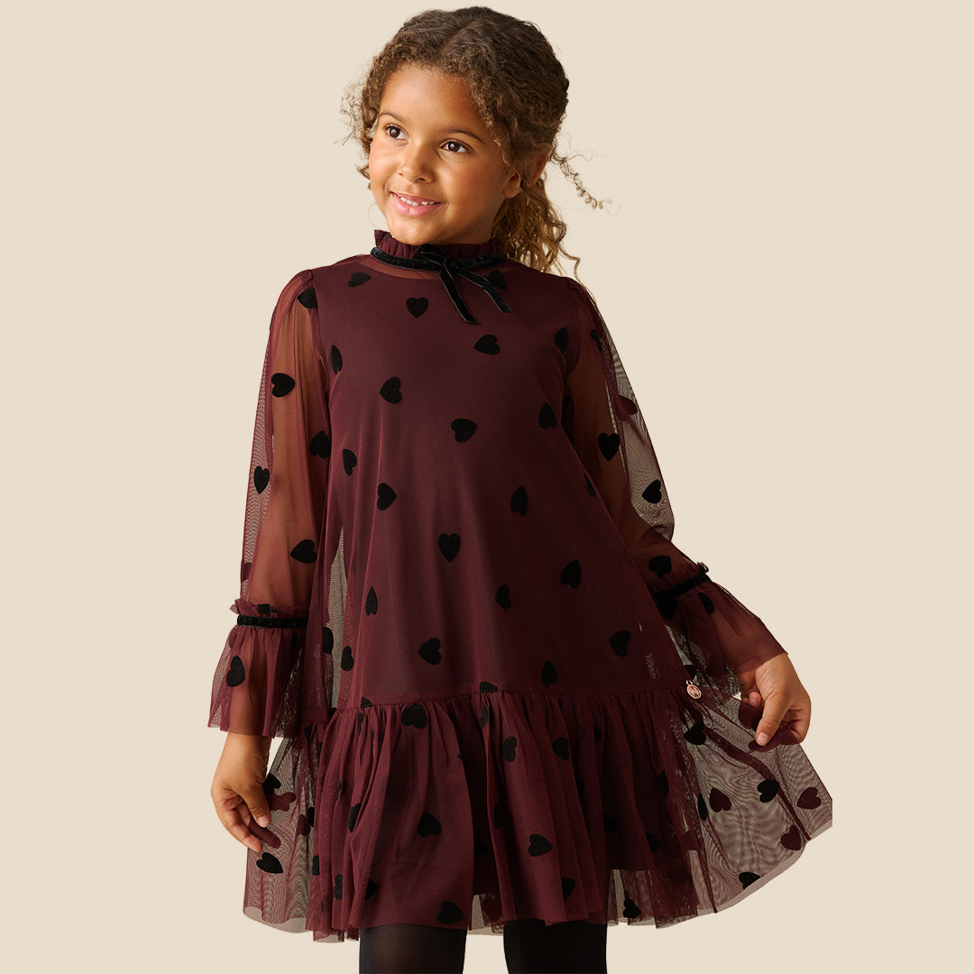 A young girl with curly brown hair wearing a burgundy sheer mesh dress with black heart patterns and a ruffled tulle hem, standing against a solid beige background.