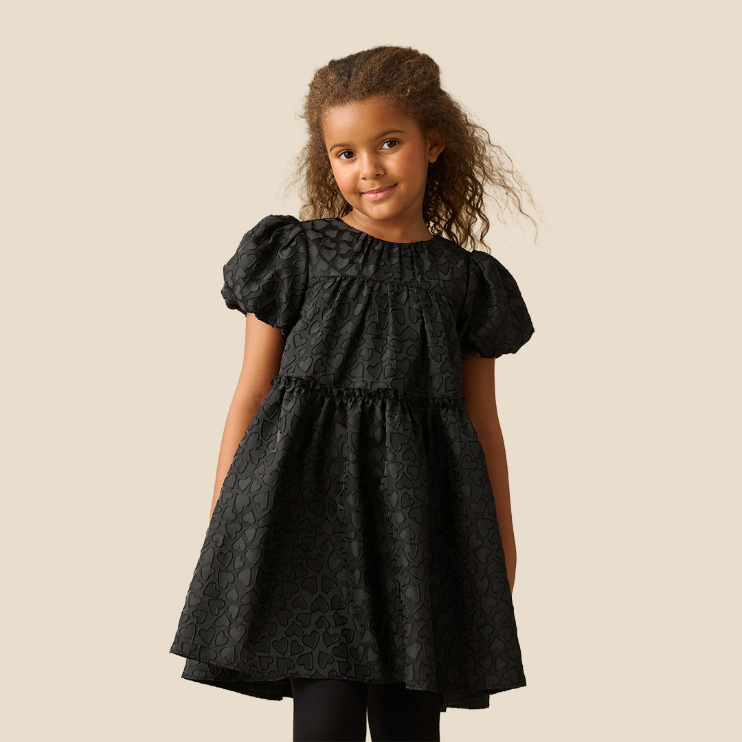 A young girl with curly brown hair wearing a black short-sleeve dress with a textured pattern, photographed against a solid beige background.