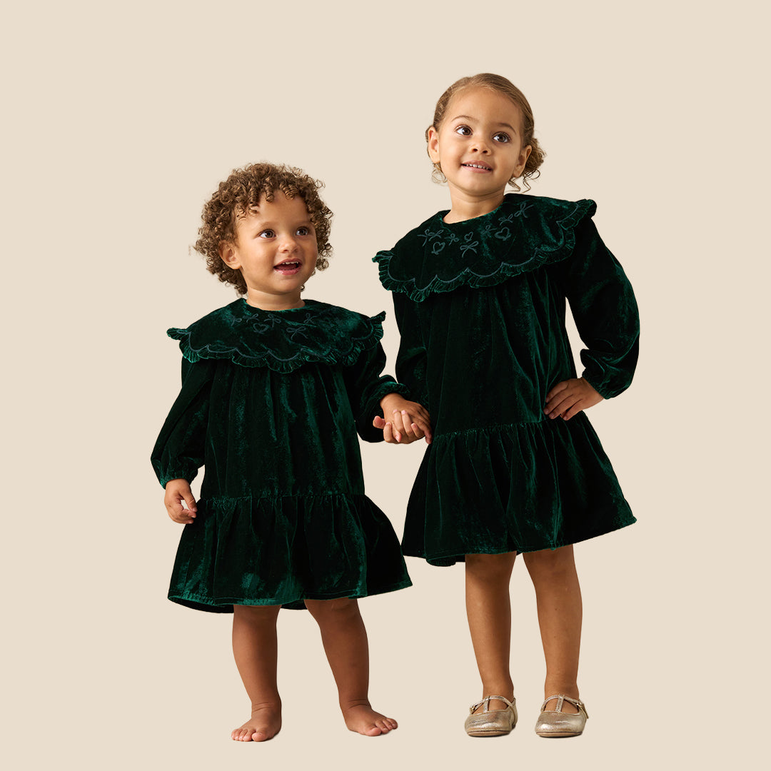 Two young children standing barefoot against a plain beige studio background, wearing matching dark green velvet dresses with wide ruffled shoulders and long sleeves.