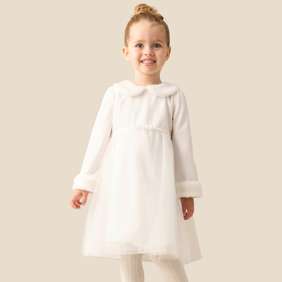 A young girl with blonde hair in a bun wearing a white long-sleeve tulle dress with faux fur trim on the collar and cuffs, standing against a solid beige background.