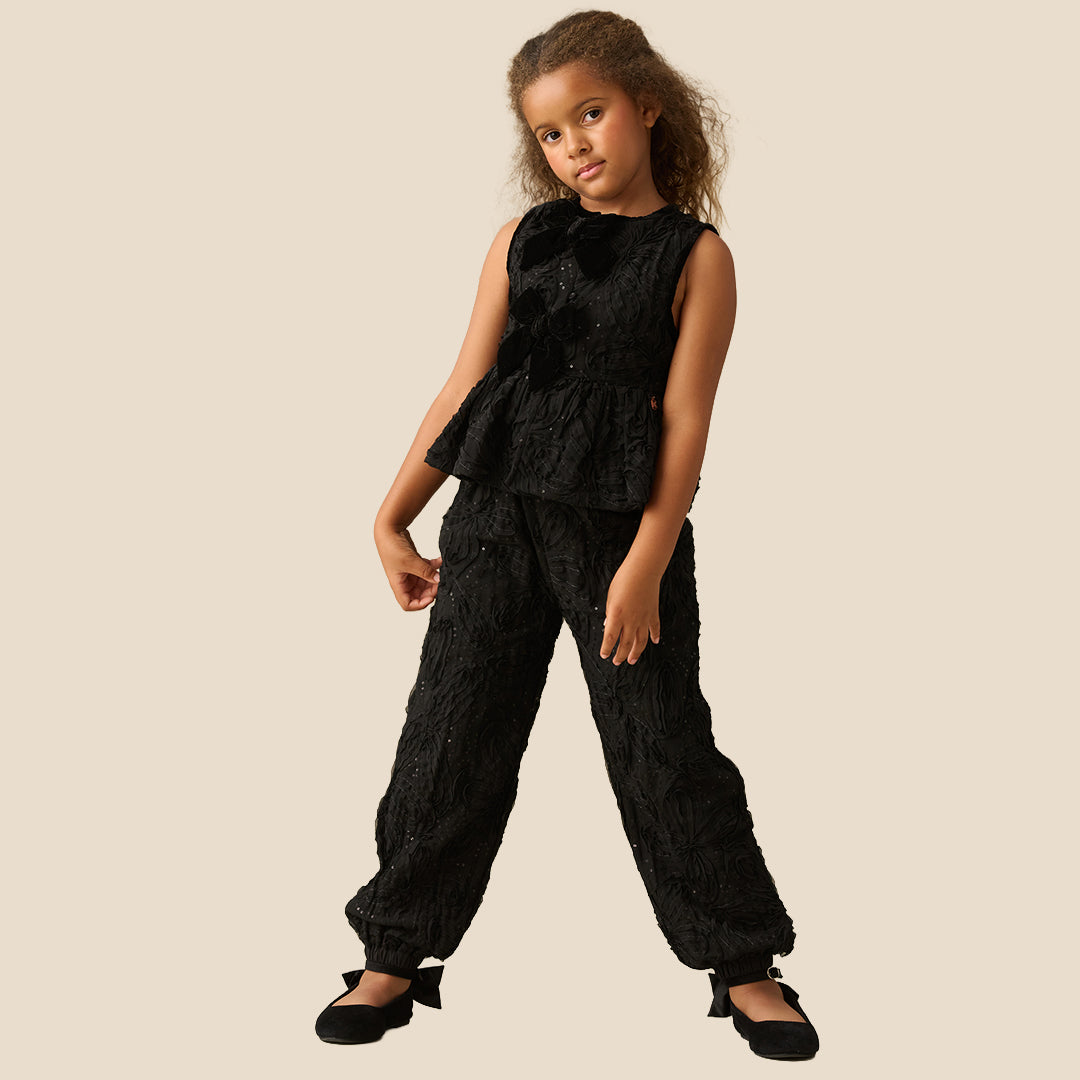 A young girl with curly brown hair wearing a sleeveless black textured dress with a ruffled peplum and bow detail, posing against a solid beige background.