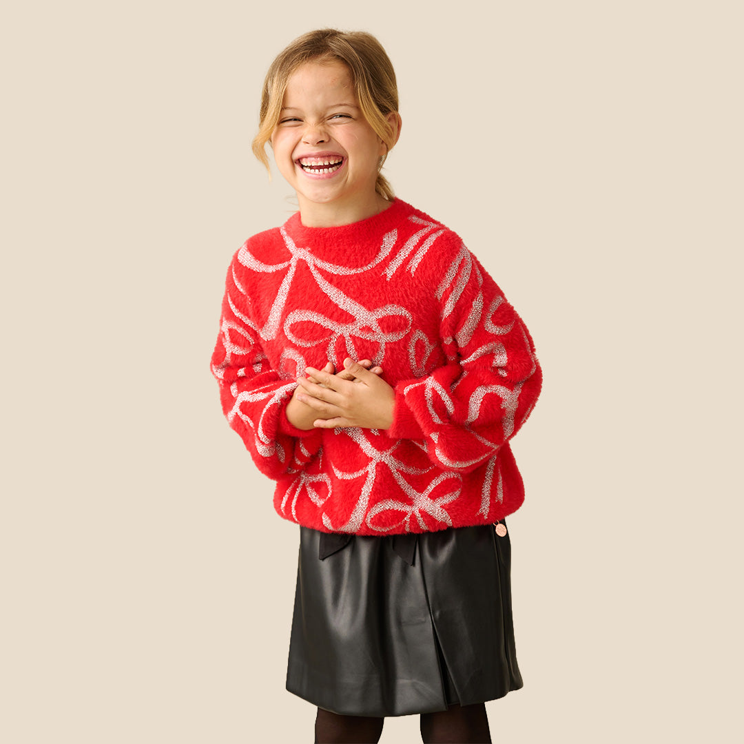A young girl smiling while wearing a bright red fuzzy knit sweater with a white abstract pattern, paired with black bottoms, against a solid beige background.