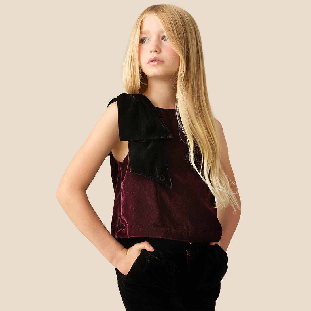 A young girl with long blonde hair wearing a sleeveless deep burgundy velvet top with a large black bow detail on the shoulder, paired with black pants, against a solid beige background.