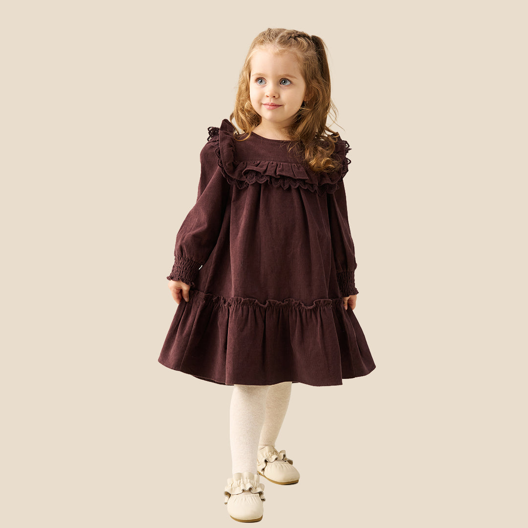 A young girl with light brown hair in pigtails stands against a solid beige background wearing a long-sleeved dark plum-brown dress with ruffles, white tights, and cream-colored shoes.