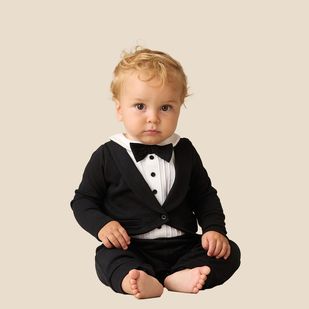 A baby wearing a black formal tuxedo-style romper with a white shirt and black bow tie, posing with arms raised against a plain beige background.