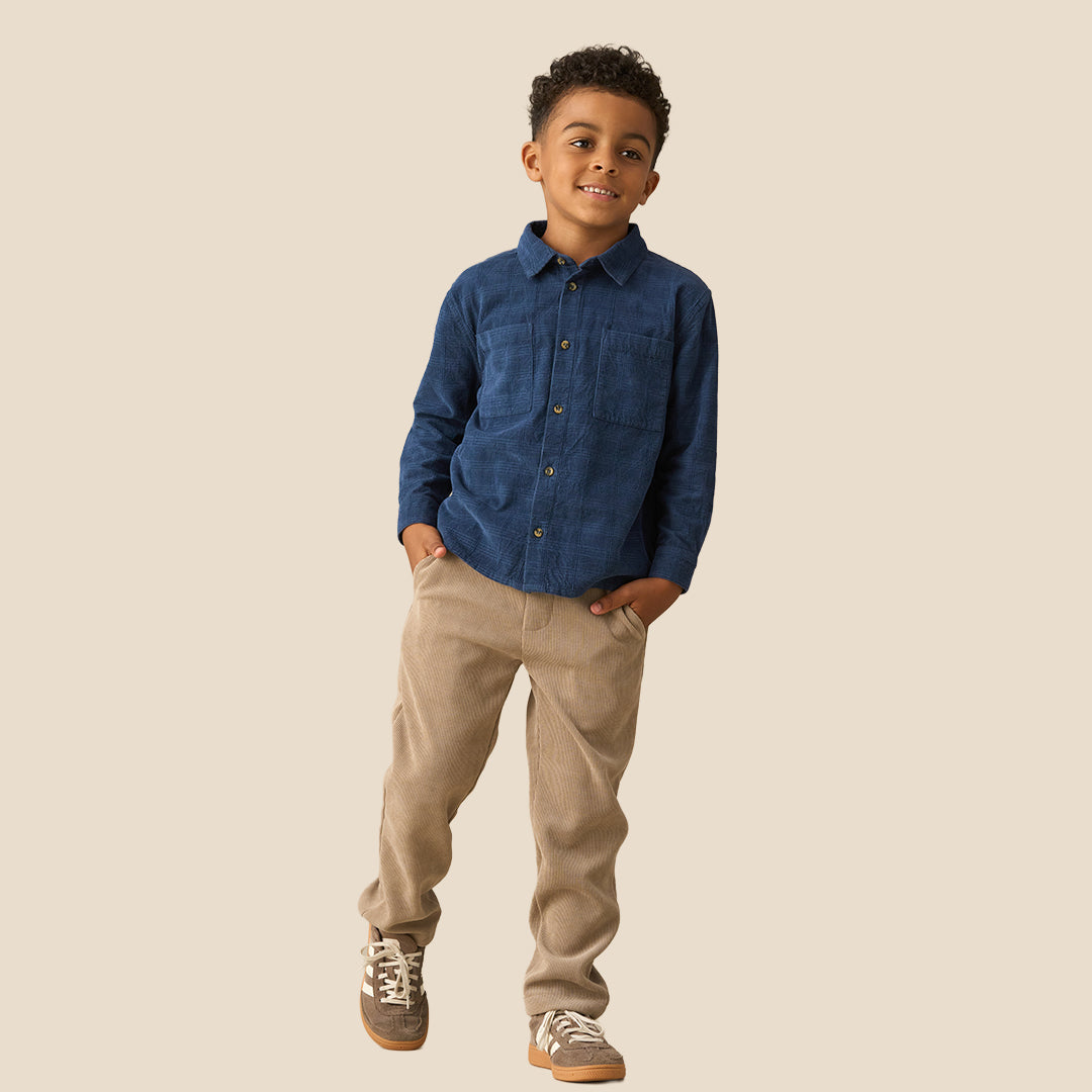 A young boy with curly dark hair wearing a dark blue long-sleeve button-up shirt and khaki pants, standing against a solid beige background.