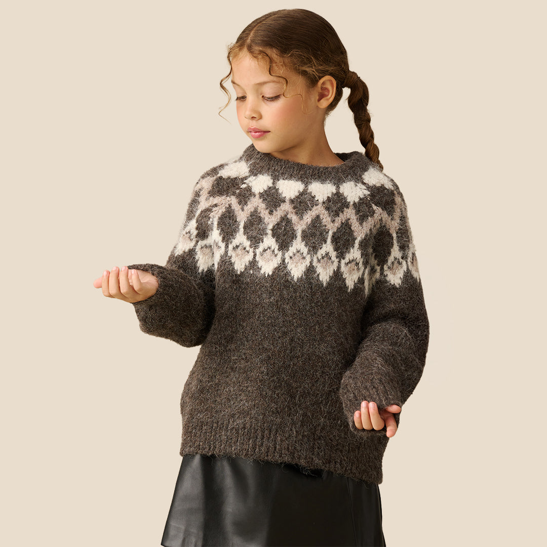 A young girl modeling a chunky knit crewneck sweater with a brown and cream argyle pattern, paired with a black faux-leather mini skirt, against a solid beige background.