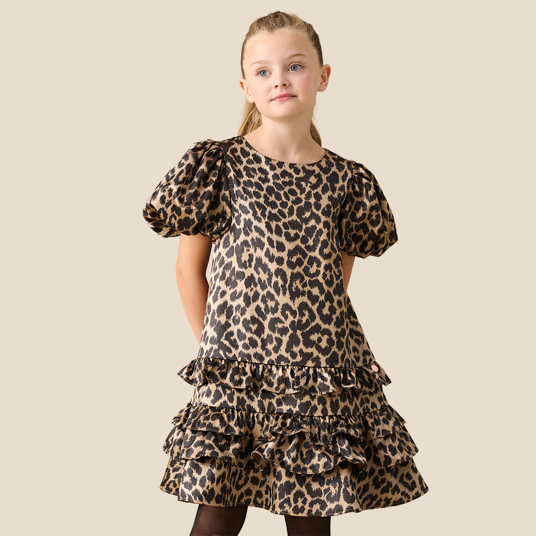 A young girl with blonde hair tied back wears a leopard print dress with puff sleeves and a tiered ruffle hem, standing against a solid beige background.