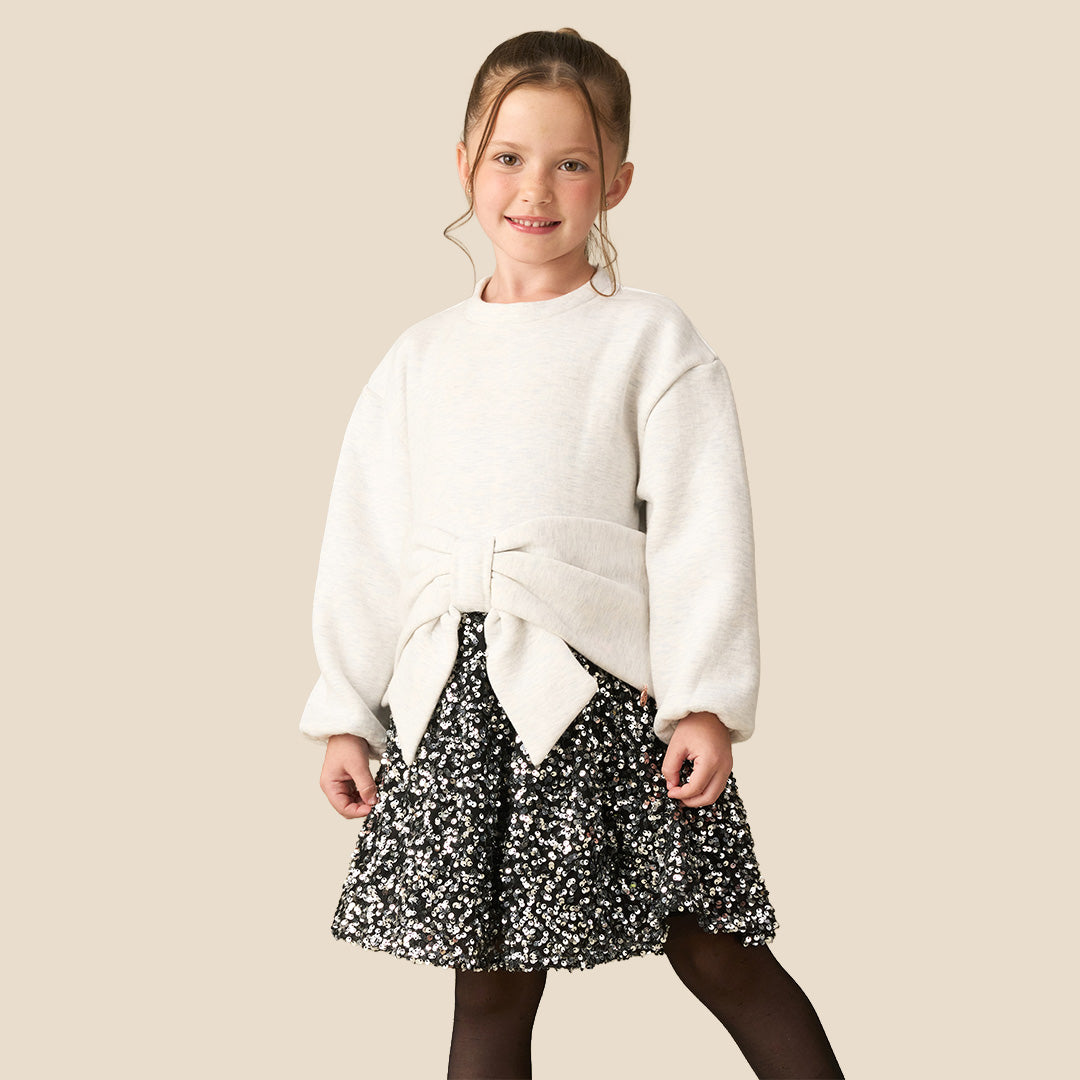 A young girl modeling a cream-colored knit sweater with a large matching fabric bow at the waist, paired with a black and white floral patterned skirt, against a solid beige background.