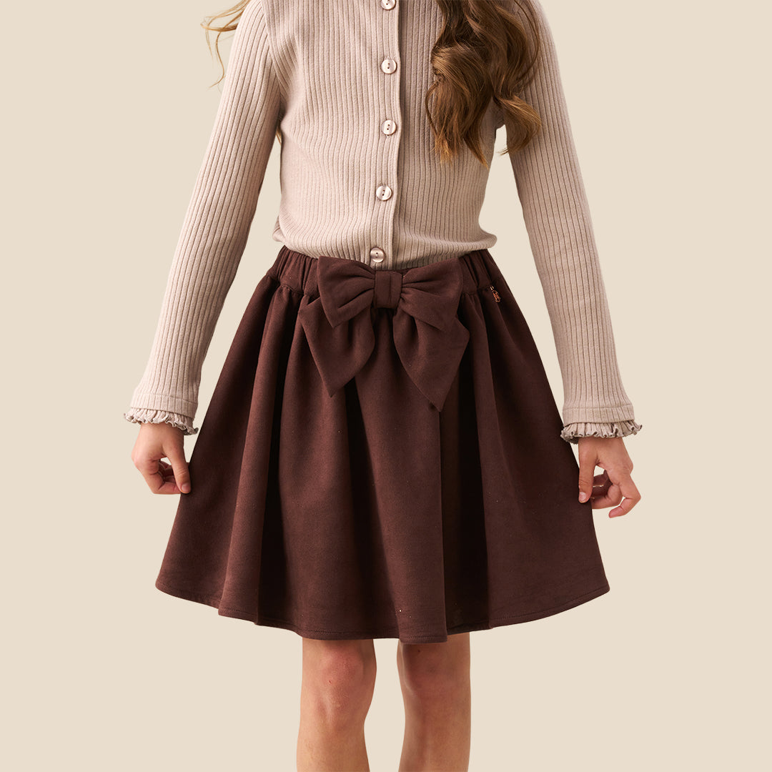 A model wearing a beige ribbed cardigan and a dark brown pleated skirt with a large bow at the waist, shown from the chest down against a solid beige background.