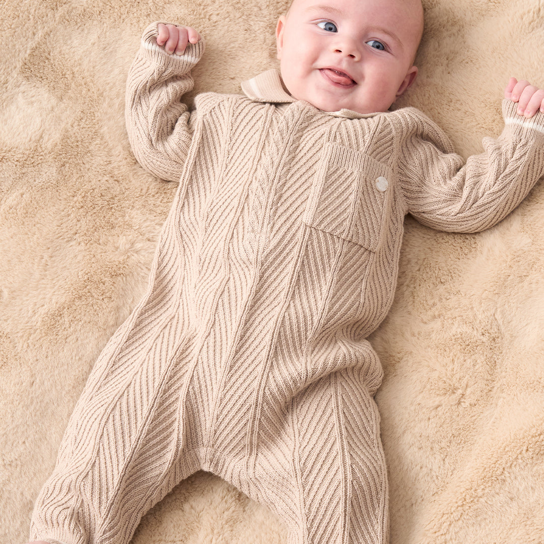 A baby wearing a beige and white textured knit one-piece jumpsuit with white shoes, standing against a solid beige background.