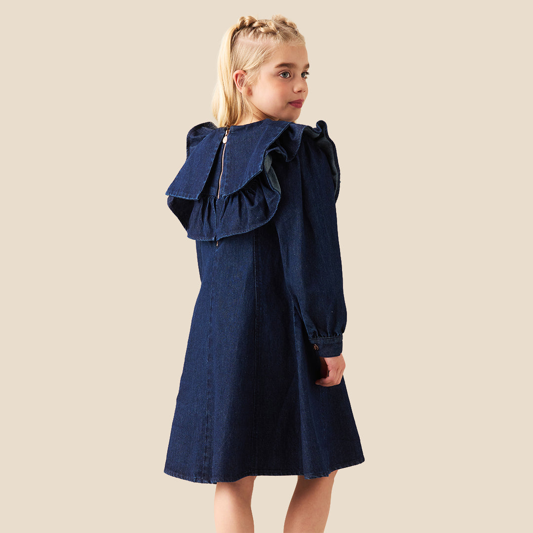 A young girl with blonde hair in two buns wearing a dark blue denim dress with a large ruffled collar, standing against a solid beige background.
