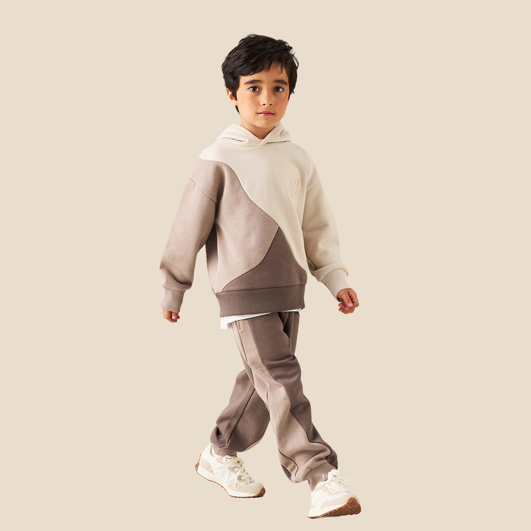 A person wearing loose taupe-brown jogger sweatpants with a white drawstring, styled with a beige and cream sweatshirt and white sneakers, against a solid beige background.
