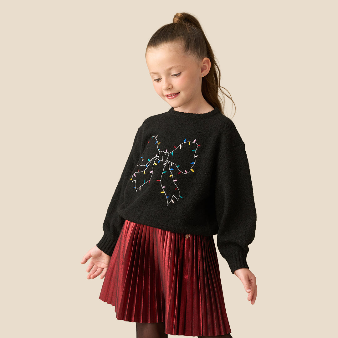 A young girl with her hair in a ponytail is wearing a black knit sweater featuring a large bow design outlined with colorful Christmas string lights, paired with a red pleated skirt, set against a solid beige background.
