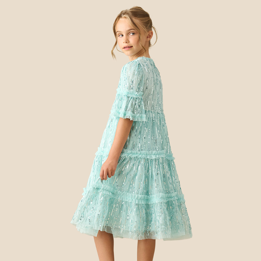 A young girl with a long brown ponytail wearing a light mint green sequined tiered dress, standing against a solid beige background.