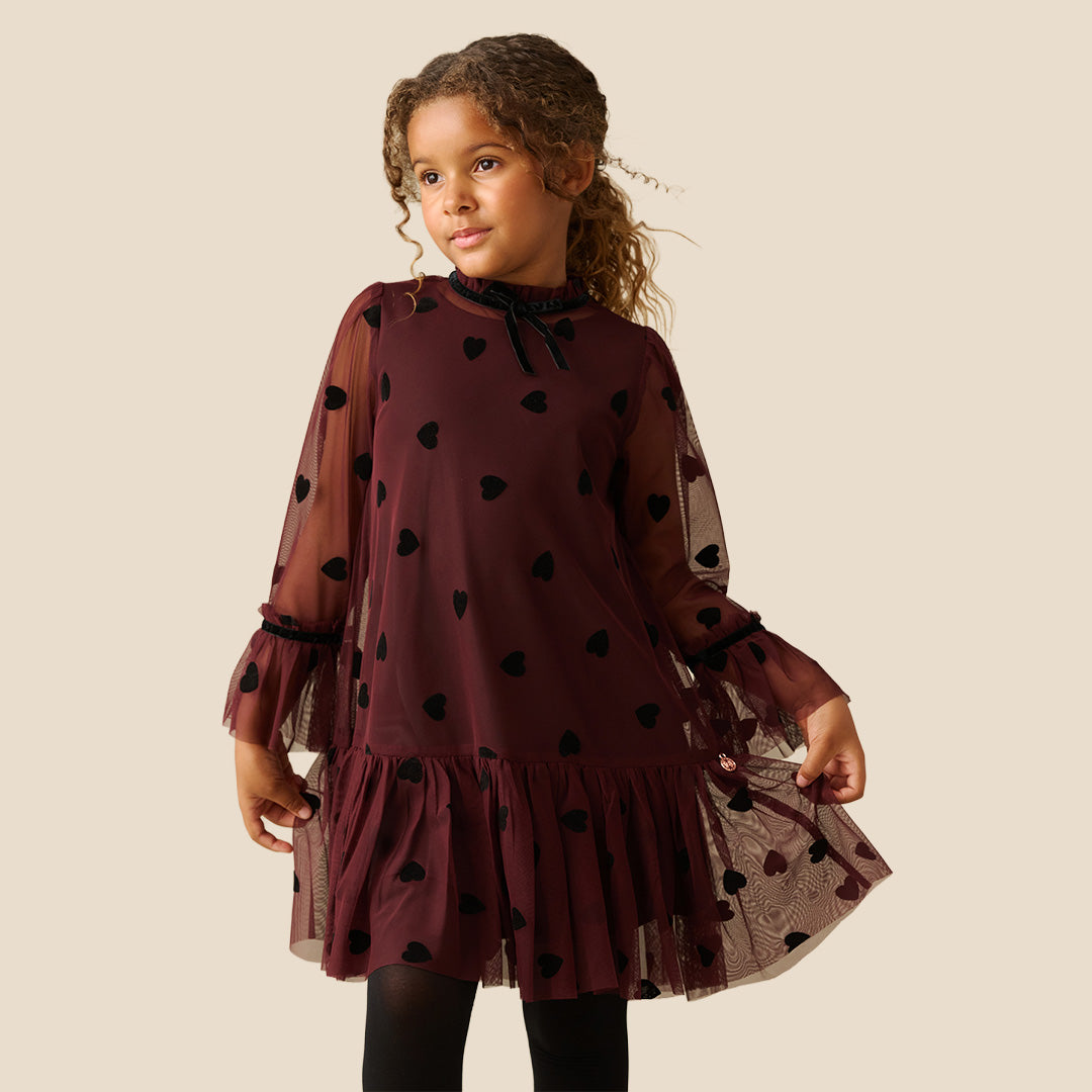 A young girl with curly brown hair wearing a burgundy sheer mesh dress with black heart patterns and a ruffled tulle hem, standing against a solid beige background.