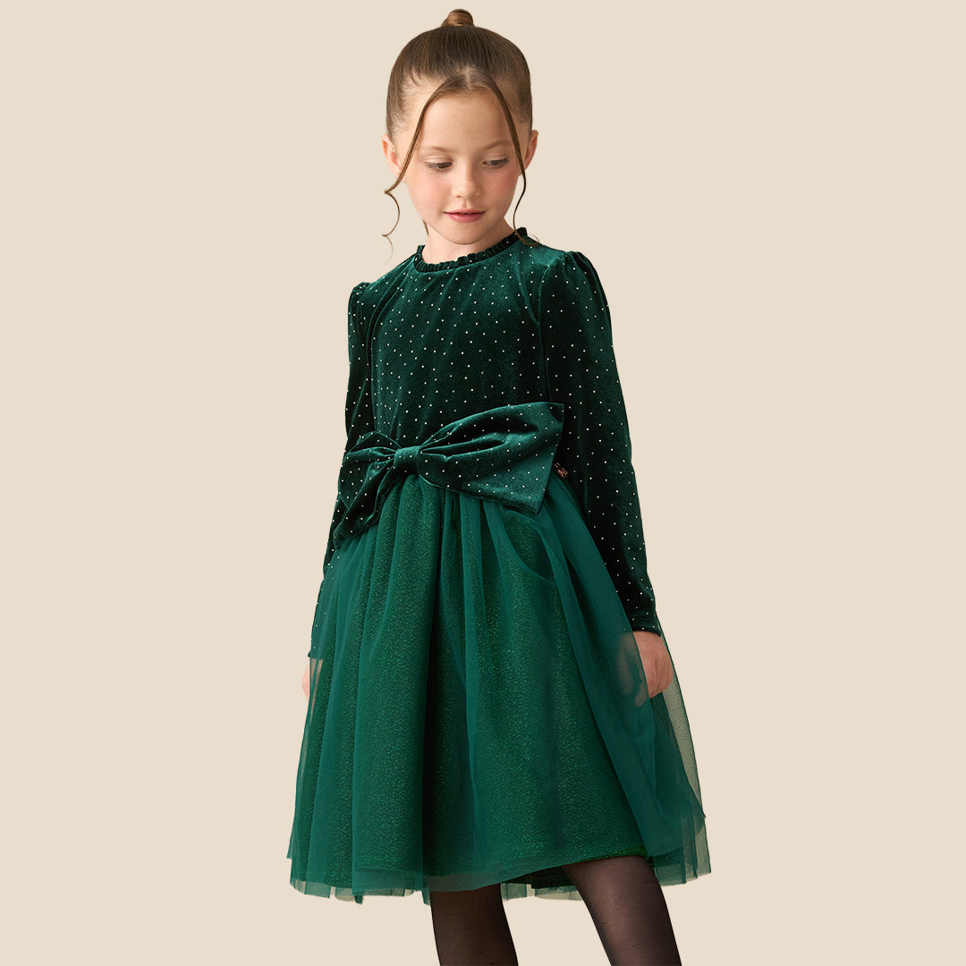 A young girl wearing a dark green velvet long-sleeve dress with a polka-dot pattern, a matching tulle skirt, and a large bow at the waist, standing against a solid beige background.