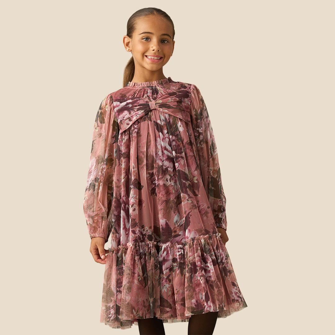 A young girl with her hair in a ponytail models a long-sleeve tiered dress with a dusty rose and brown floral print against a solid beige background.