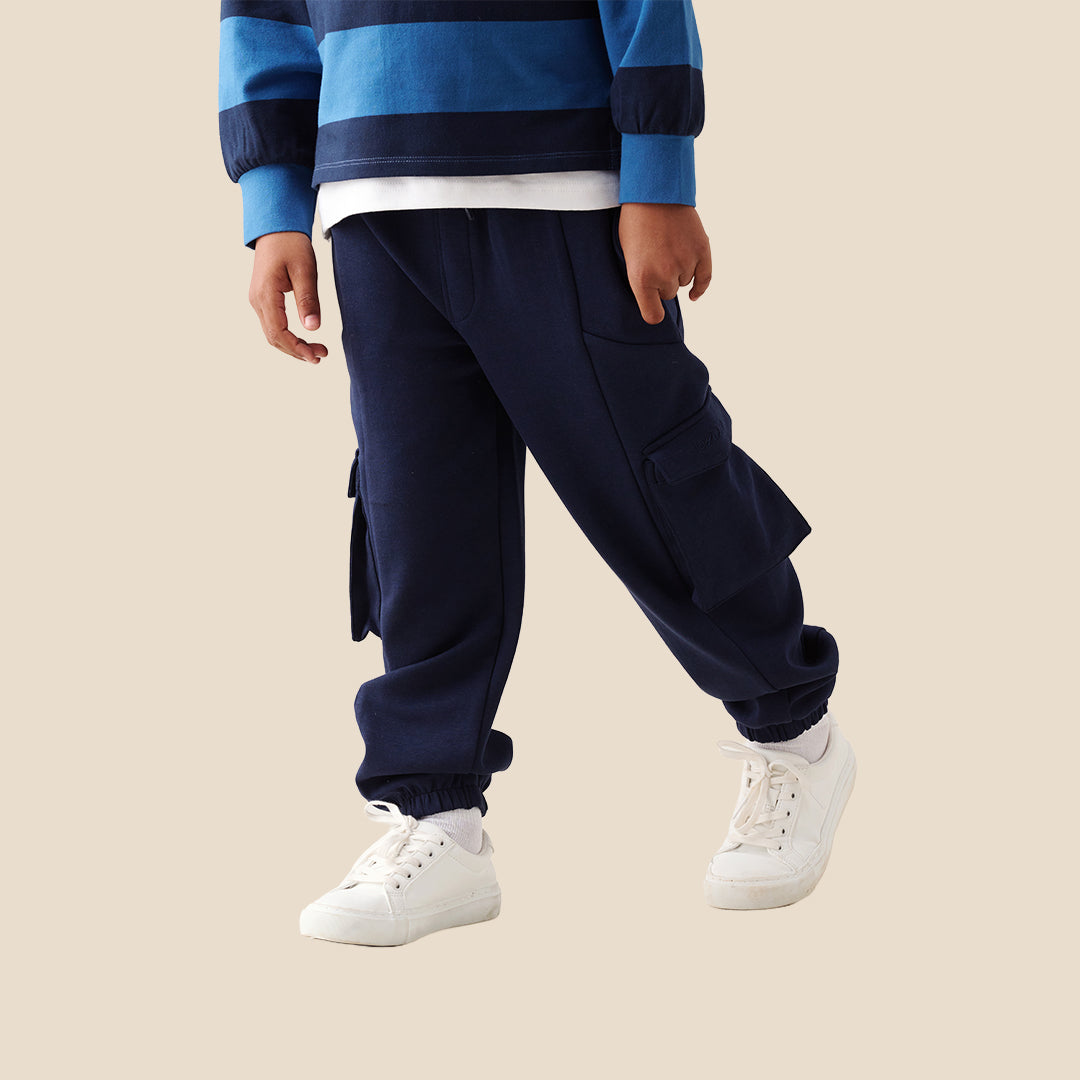 A child wearing navy blue cargo pants with side flap pockets, a blue and navy striped long-sleeve shirt, and white sneakers, standing against a solid beige background.