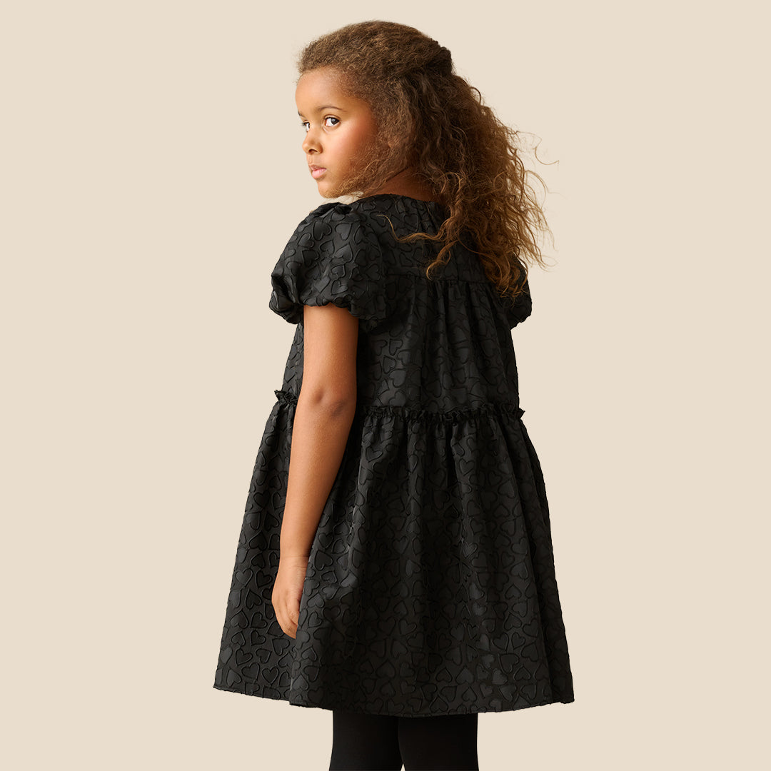 A young girl with curly brown hair wearing a black short-sleeve dress with a textured pattern, photographed against a solid beige background.