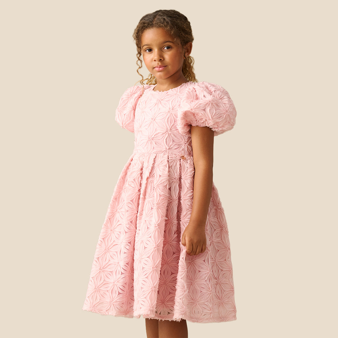A young girl with curly brown hair wearing a light pink textured dress with puffed short sleeves and a full skirt, standing against a solid beige background.