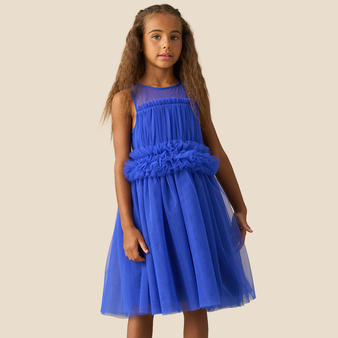 A young girl with long brown hair wearing a sleeveless royal blue tulle dress with a ruffled waist, standing against a solid beige background.