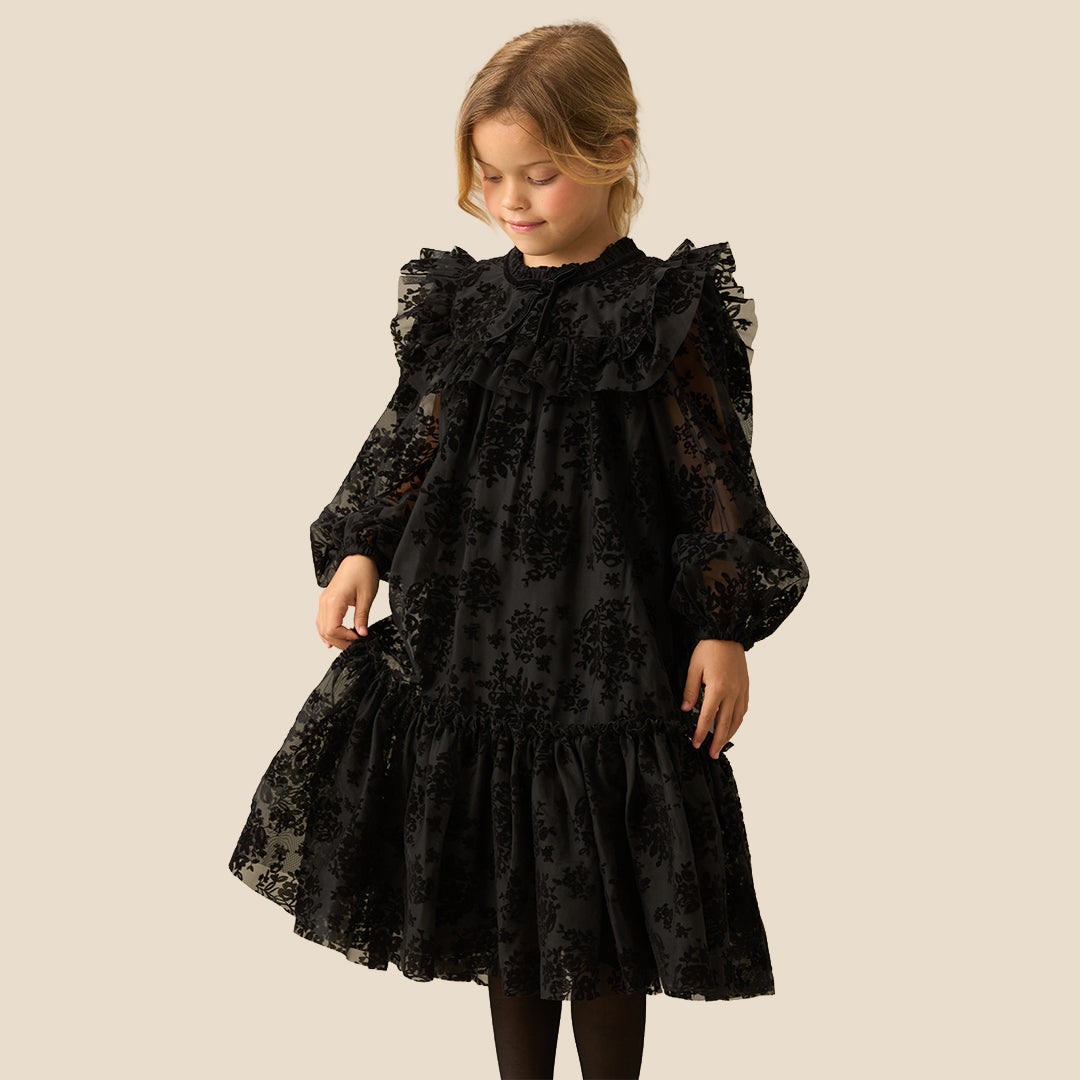 A young girl with blonde hair smiling while wearing a black lace long-sleeve dress with ruffled details, photographed against a solid light beige background.