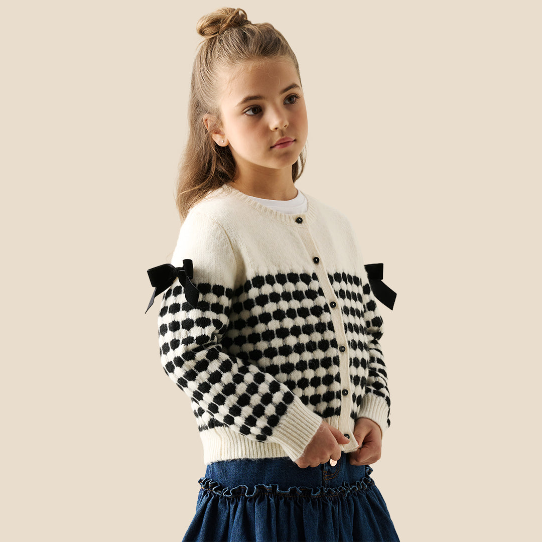 A young girl with her hair in a bun wears a cream knit cardigan with black polka-dot patterned sleeves and black ribbon bows on the shoulders, paired with a dark blue ruffled skirt, against a solid beige background.