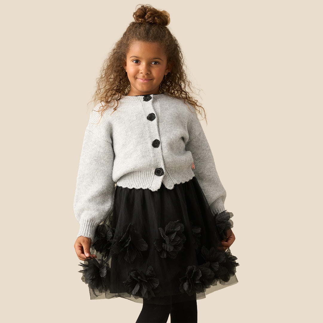 A young girl with curly brown hair wearing a light gray knit cardigan with black buttons and a black ruffled skirt, standing against a solid beige background.