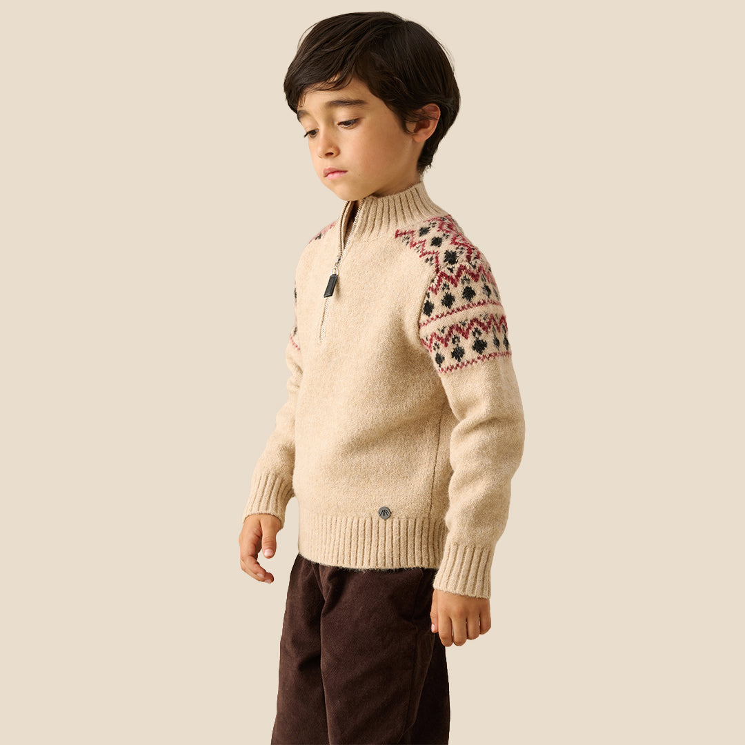 A young boy wearing a beige knit half-zip sweater with red and black Fair Isle patterns on the shoulders and sleeves, set against a solid light beige background.