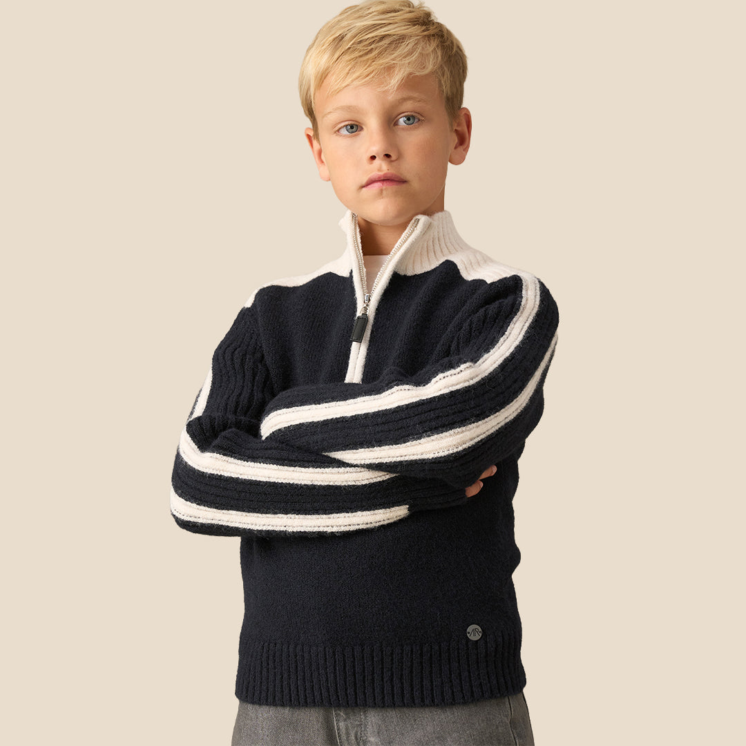 A young boy wearing a navy blue and cream knit quarter-zip sweater with white stripe details on the sleeves, paired with gray pants, against a solid beige background.