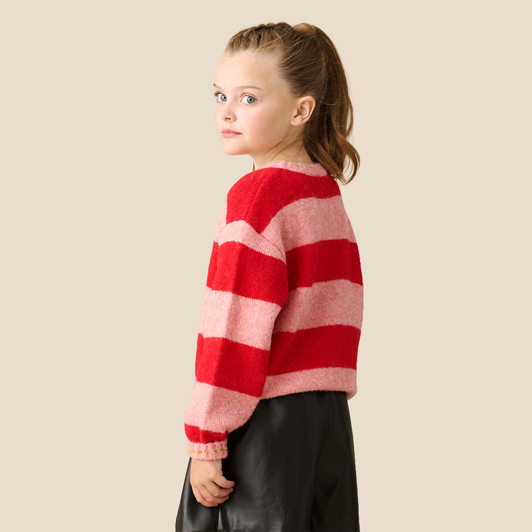 A young girl wearing a fuzzy knit sweater with red and pink horizontal stripes, featuring 'Merry Night' in gold glitter script across the chest, paired with a black faux-leather mini skirt, set against a solid beige background.