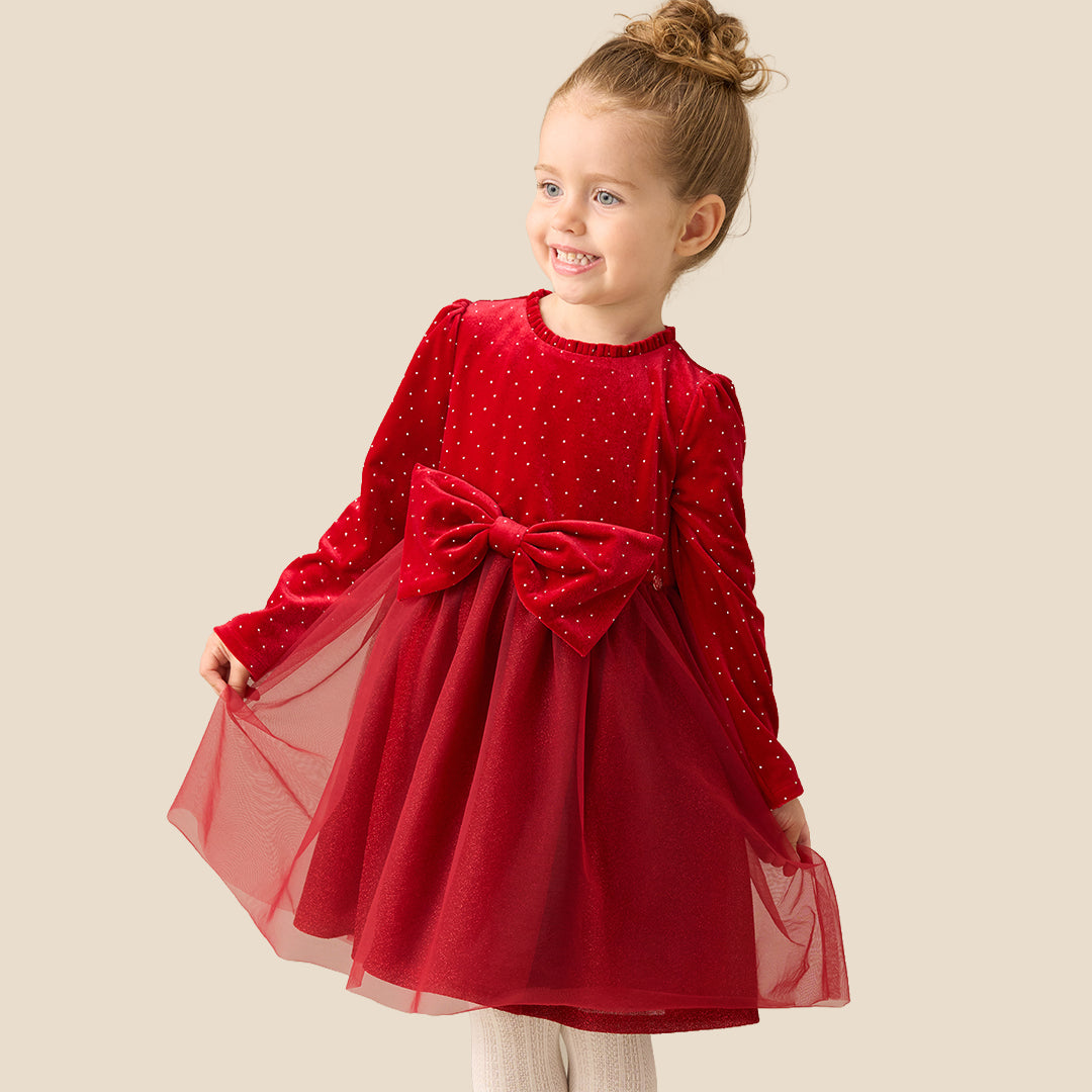 A young girl with her hair in a bun stands against a solid beige background wearing a festive red dress with a polka-dot velvet bodice, long sleeves, a large matching bow at the waist, and a tulle skirt. She is also wearing white knit tights and metallic gold shoes.