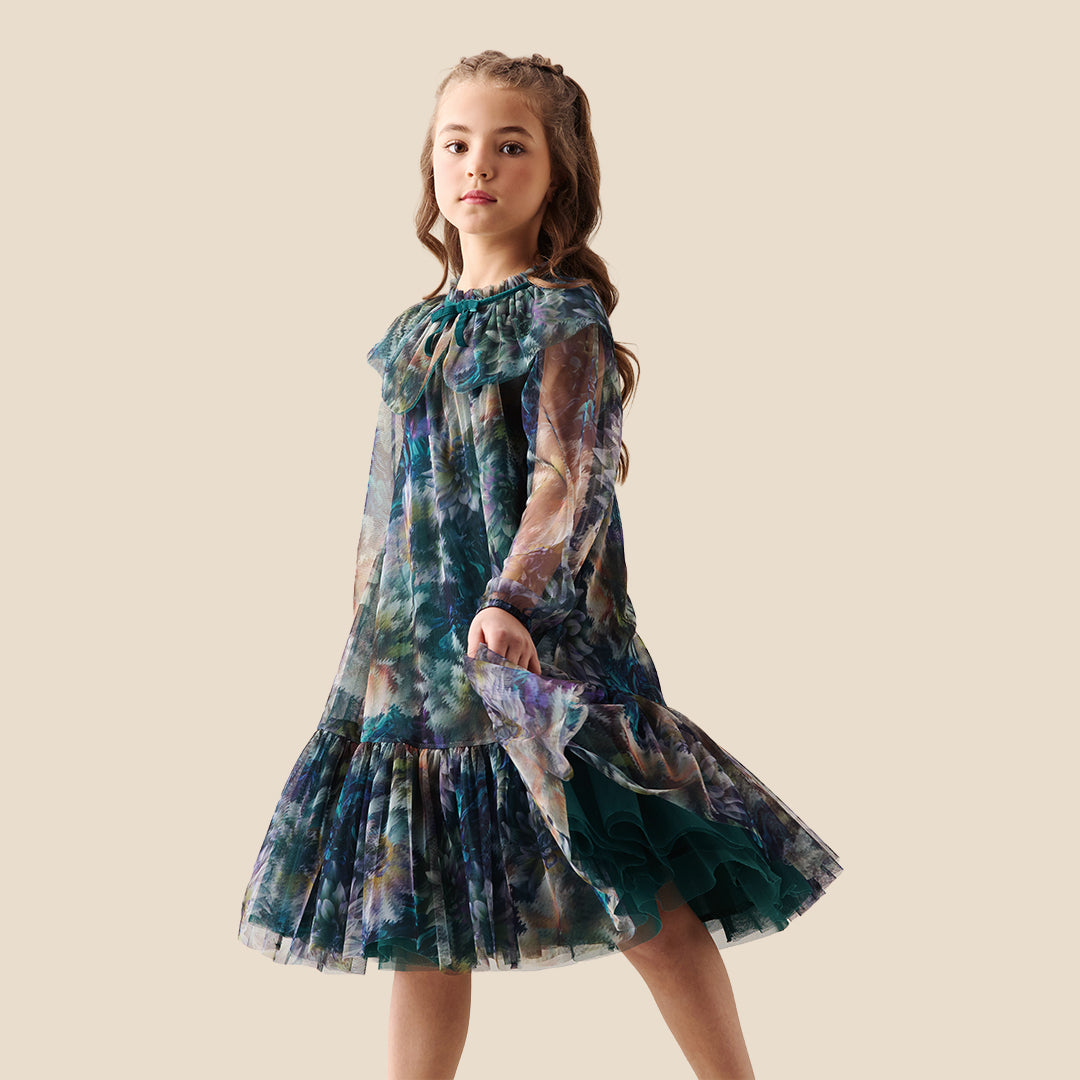 A young girl modeling a sheer, tiered tulle dress with a large ruffled collar and a blue, green, and purple floral watercolor pattern, set against a solid beige background.