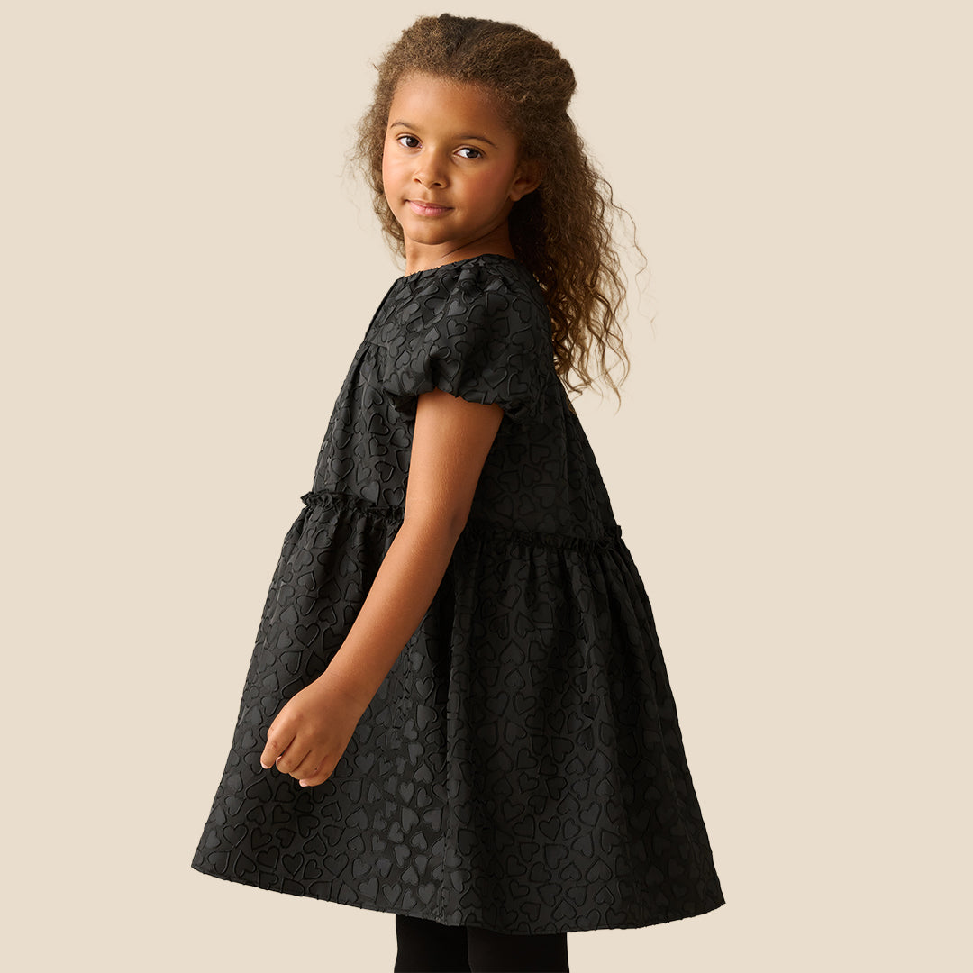 A young girl with curly brown hair wearing a black short-sleeve dress with a textured pattern, photographed against a solid beige background.